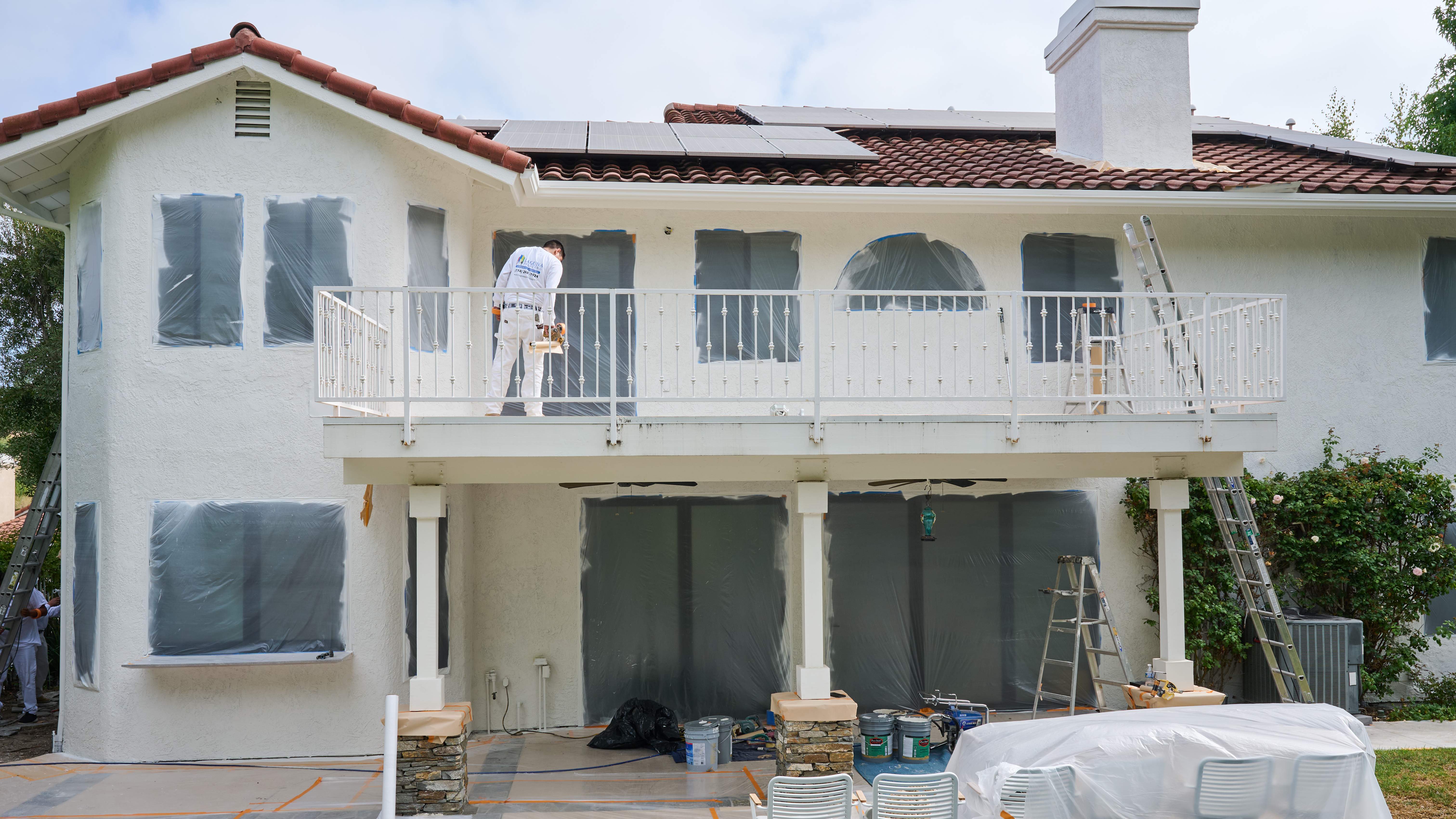 Home exterior prepared for painting with windows and furniture covered as a painter works on the balcony